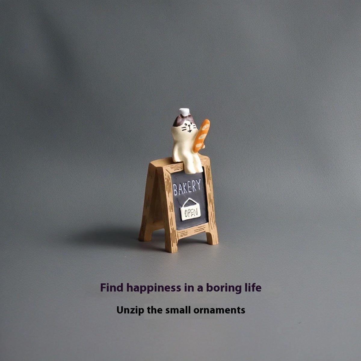 Cat figurine holding baguette and sitting on small BAKERY OPEN chalkboard sign – Pausadesk