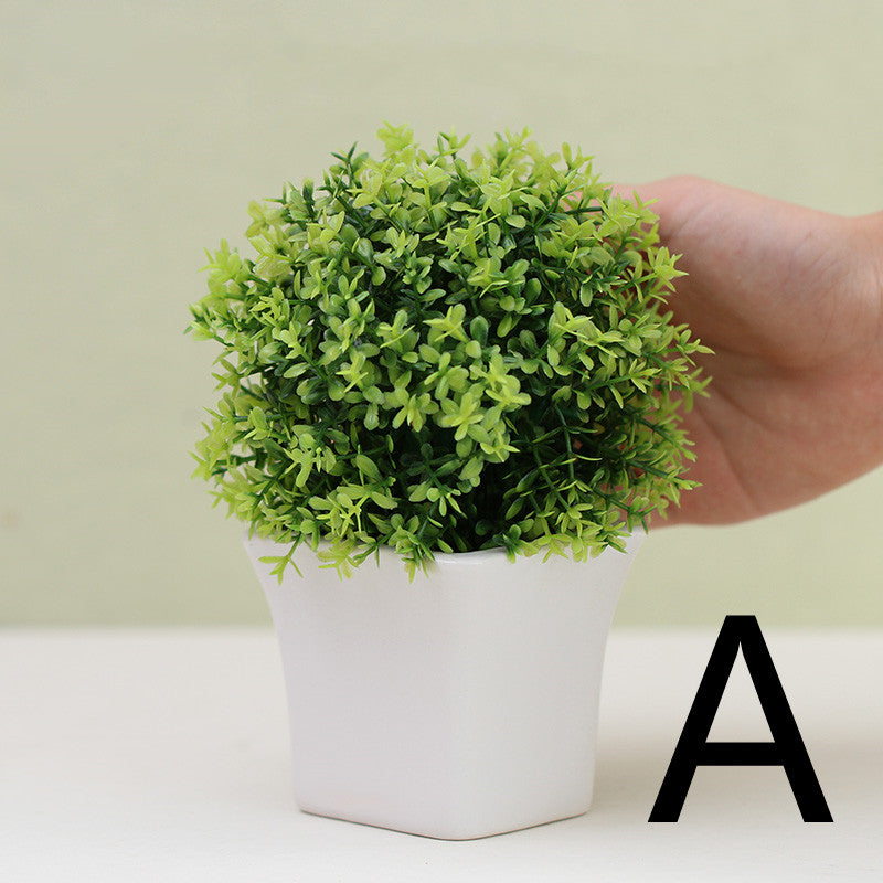 Green artificial mini plant in white square pot, held by hand for scale – Pausadesk