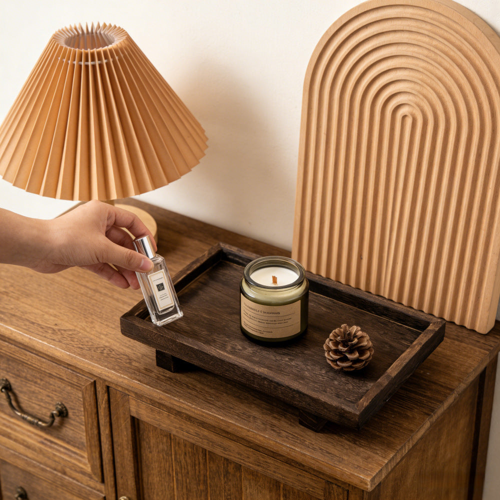 Wooden tray with candle and pinecone on a dresser, hand adding a perfume bottle beside it – Pausadesk